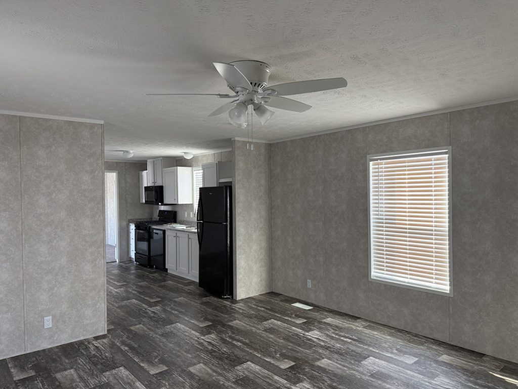 A modern, unfurnished room with gray walls, dark wood flooring, a ceiling fan, and a window with blinds offers a contemporary vibe. The space features a view of the kitchen area with white cabinets and a black refrigerator.