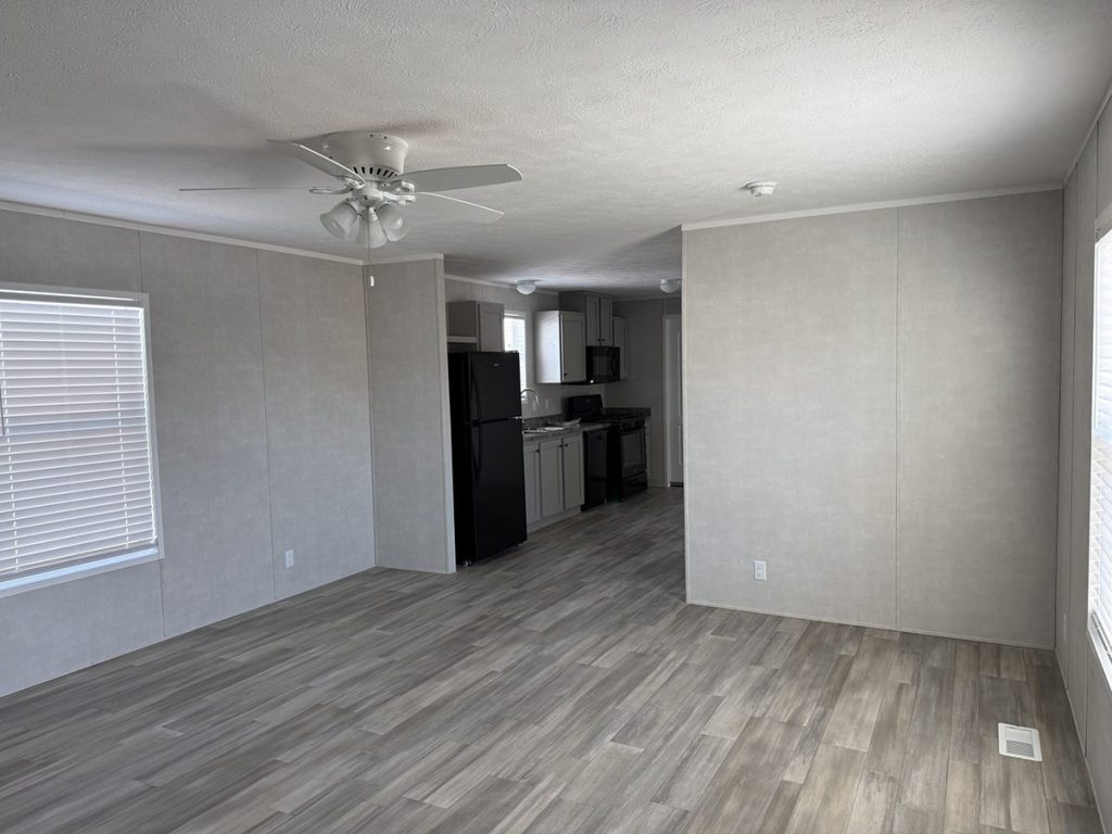 A modern, empty room with light wood flooring, gray walls, a ceiling fan, and large windows awaits you. The kitchen with black appliances and gray cabinets is visible in the background.