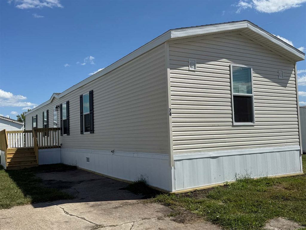 A beige mobile home with white trim, black shutters, and a small wooden porch sits on a grassy lot beside a concrete driveway under a blue sky with scattered clouds.