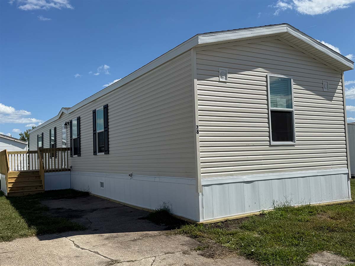 A beige mobile home with white trim, black shutters, and a small wooden porch sits on a grassy lot beside a concrete driveway under a blue sky with scattered clouds.