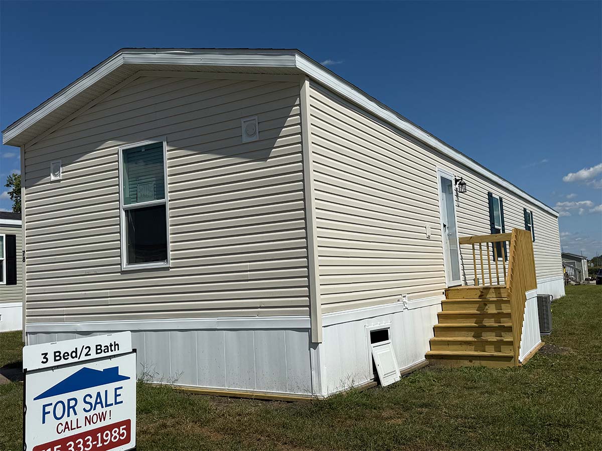 A beige mobile home with white trim, a small wooden porch, and a "For Sale" sign in the yard. The sign indicates 3 bedrooms, 2 baths, and a phone number to call. The sky is clear and sunny.