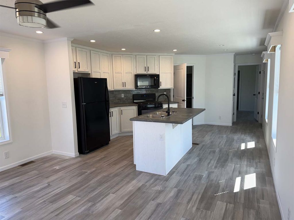 Modern kitchen with white cabinets, black appliances, and a center island. The space features gray wood-like flooring, recessed lighting, and an open layout that leads to a hallway. Natural light enters through adjacent windows.