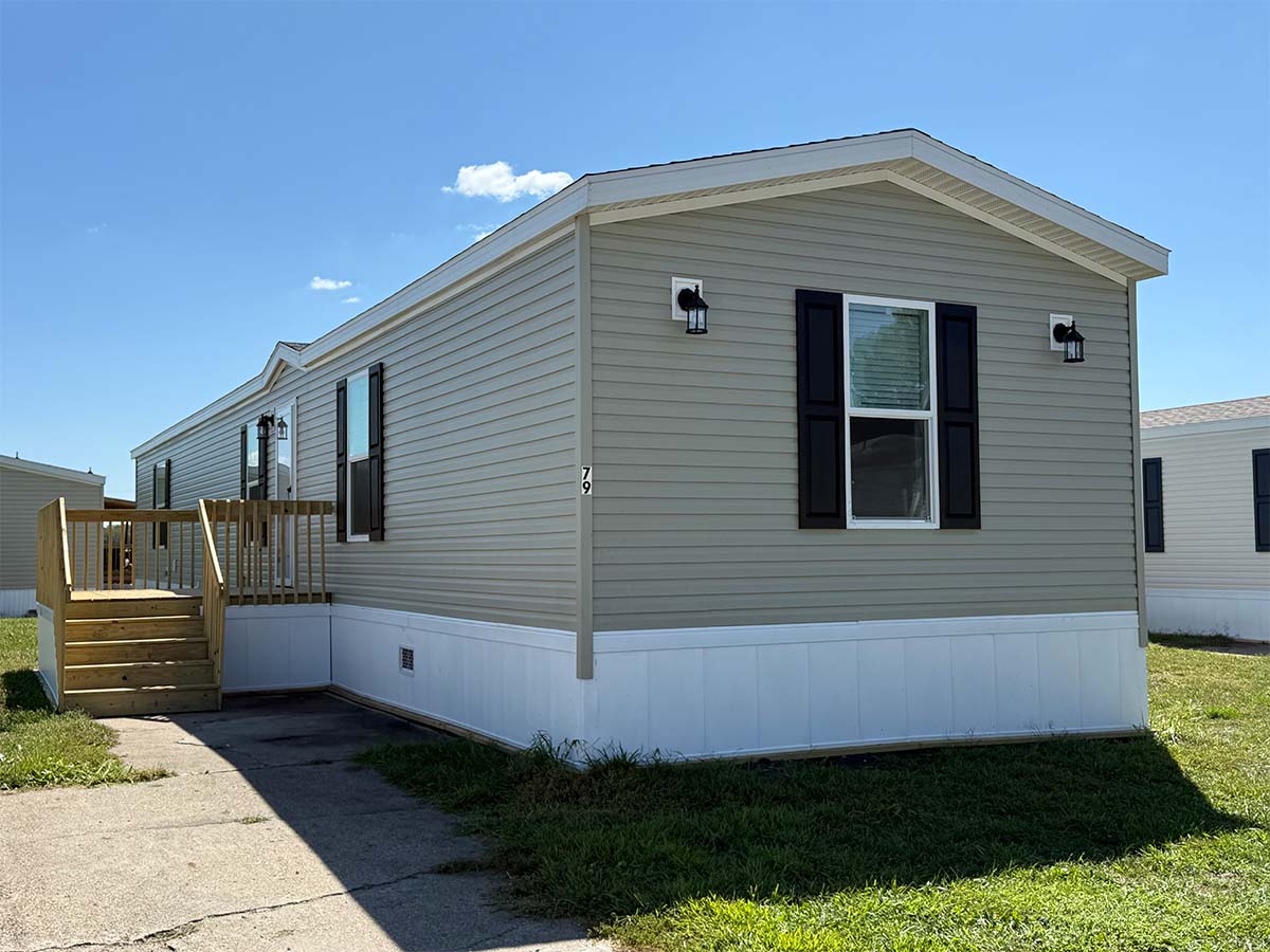 Light tan manufactured home with black shutters, two wall lights, a small wooden porch with steps, and a ramp. The home sits on grass next to a concrete walkway under a clear blue sky.