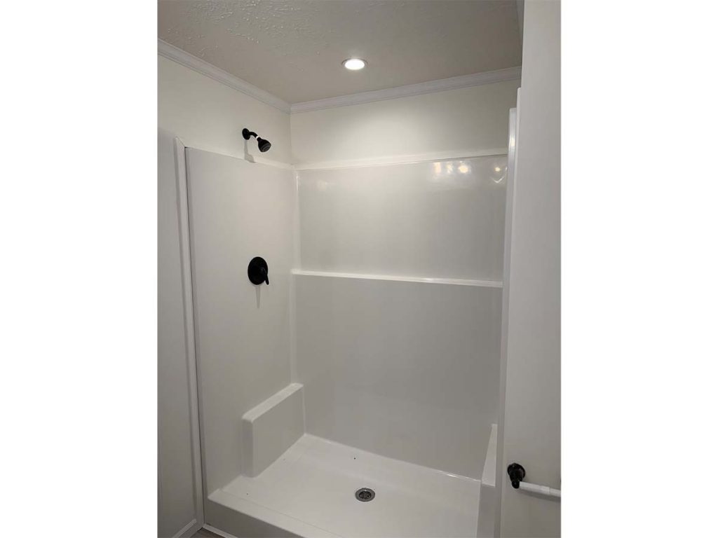 A white, modern walk-in shower with a black showerhead, a built-in seat, molded shelving, and a recessed ceiling light above. The shower has smooth walls and a drain in the center of the floor.