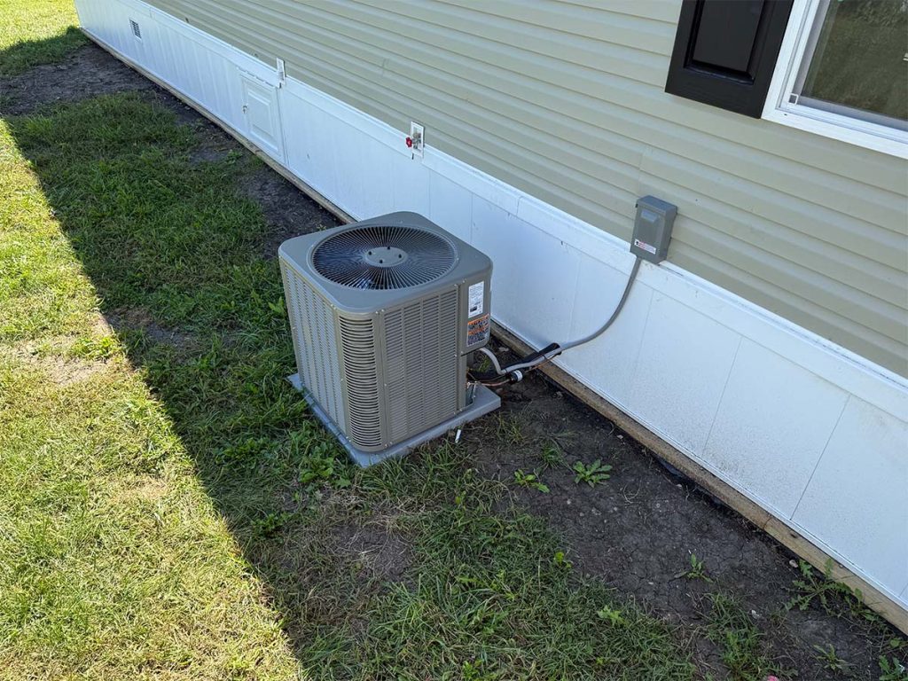 A central air conditioning unit installed outside a beige building, connected by electrical and refrigerant lines, sits on a concrete pad next to a grassy area.